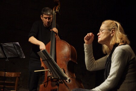 Die ältere Schauspielerin sitzt auf einem Stuhl und erzählt. sehr nachdenklich. Hinter ihr spielt der Musiker den Kontrabass.