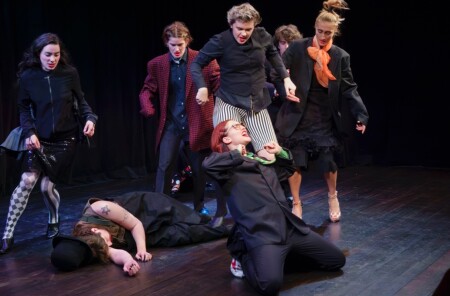 Auf der Bühne kniet eine Frau, hinter ihr liegt jemand auf dem Boden. Sie Spieler drum herum fokussieren auf die Frau