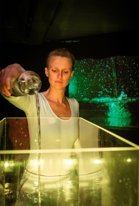 Die Performerin steht hinter einem grossen Glasbecken, das mit Wasser gefüllt ist. Sie giesst Wasser ein.