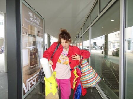 Die Frau ist nahe bei der Kamera, trägt eine rote Jacke und viel Gepäck.