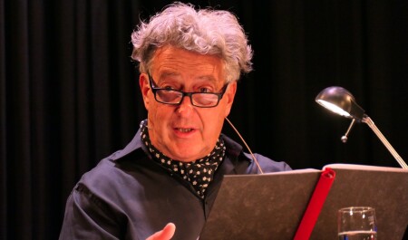 Porträt von Ernst Süss. Er steht an einem Lesepult, hat eine Brille auf und liest vor.